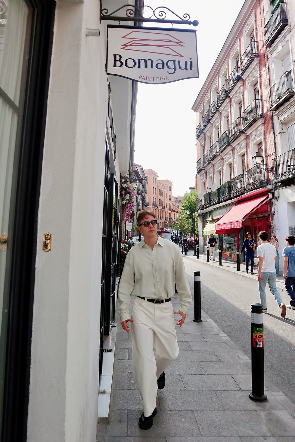 The Trendy Man con camisa clara y gafas camina por calle con edificios coloridos. Letrero de "Bomagui Papelería" visible. Día soleado.