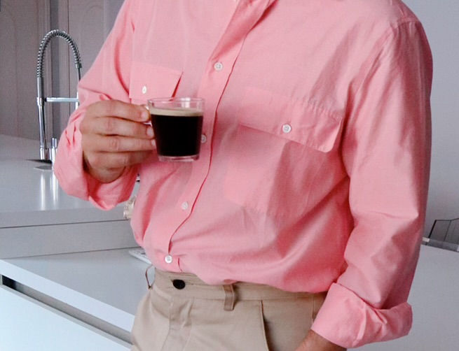 The Trendy Man con camisa rosa sostiene una taza de café oscuro en una cocina moderna junto al fregadero.