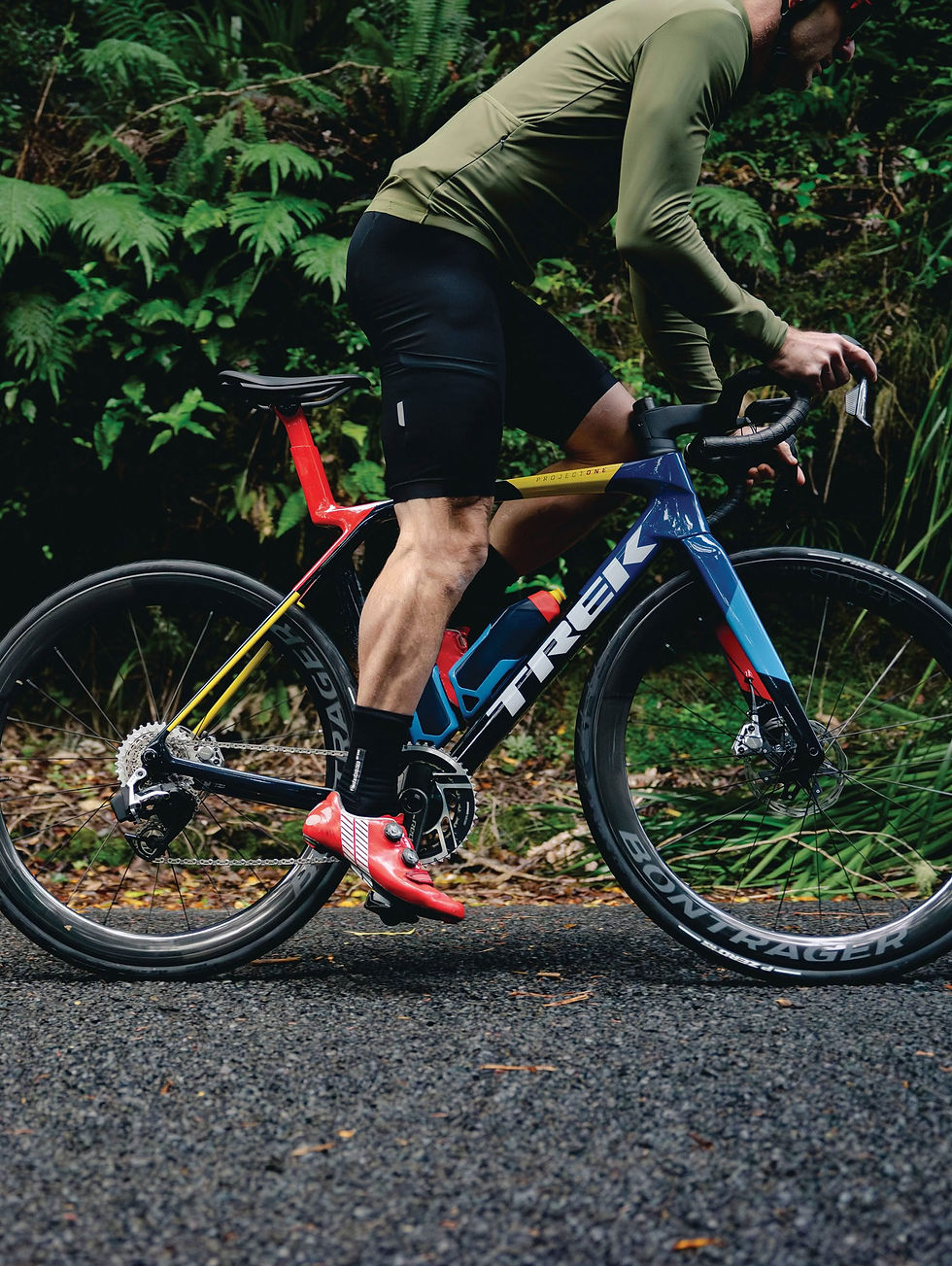 Ciclista en bicicleta Trek azul y roja en carretera de bosque, con maillot verde y zapatillas rojas, en postura de esfuerzo.