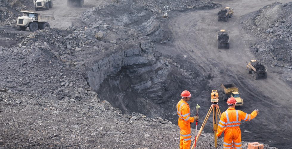 Two mine workers dressed in orange jumpsuits and wearing hardhats standing near land survey equipment near a rock mine with heavy mining trucks moving in the background