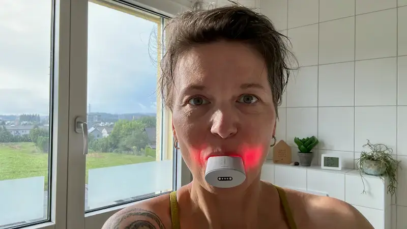 Sandra Hoppenz mit Zahnlichtanwendung: Sandra Hoppenz mit kurzen braunen Haaren und einem grünen Top lächelt in die Kamera, während sie ein Gerät mit rotem Licht im Mund hat.