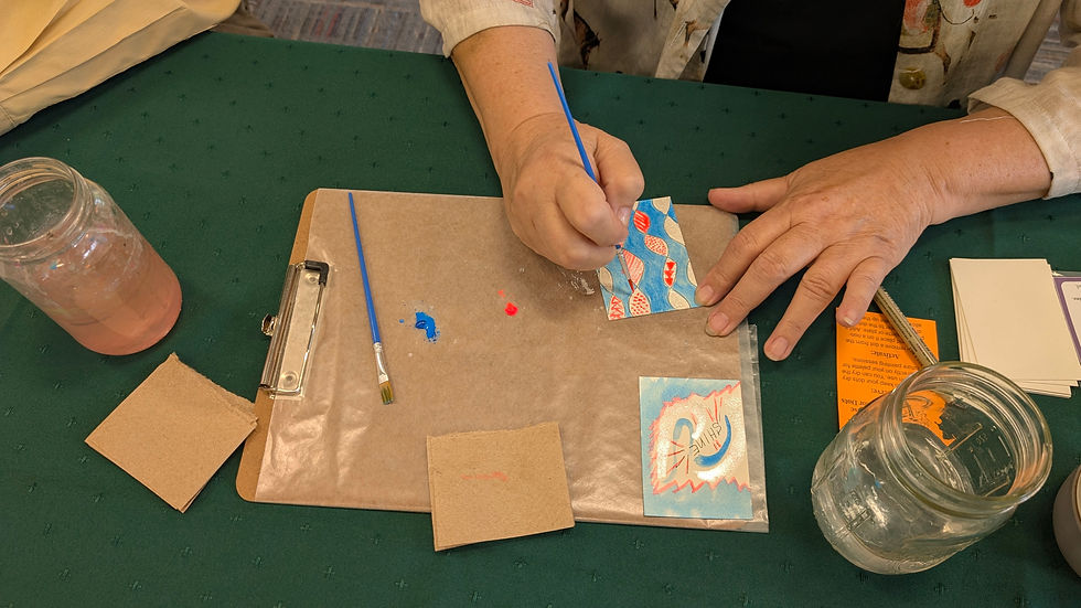 Top-down view of a workshop participant's hands painting a 2.5 by 3.5 inch art card with a fine brush, working alongside a clipboard palette with blue and pink watercolor droplets and a completed card that reads "SHINE."
