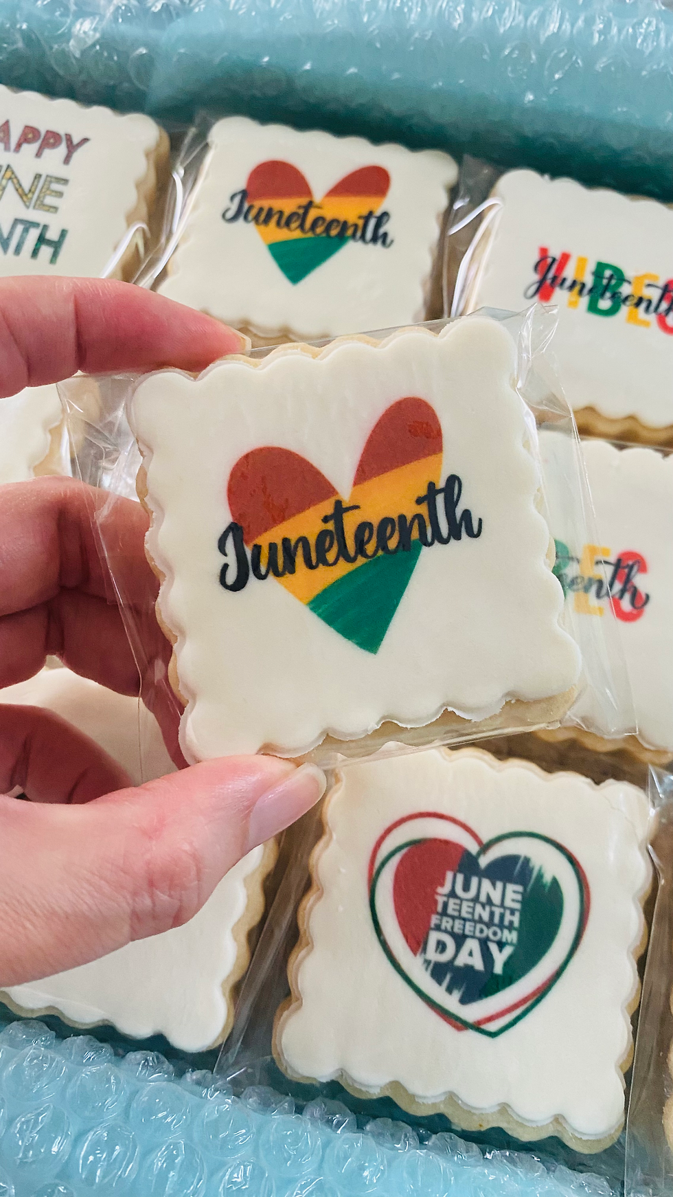 Hand holding a decorated cookie with a heart and "Juneteenth" text. Other themed cookies in the background, set against bubble wrap.