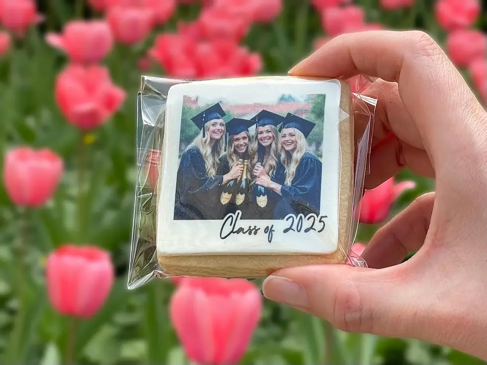 Hand holds a cookie with a photo of three graduates in caps and gowns. "Class of 2025" text. Pink tulip field in background.