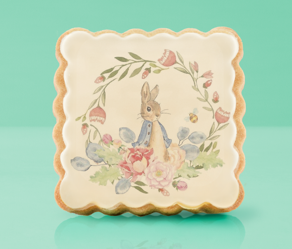 Illustrated cookie on green background, features Peter Rabbit in a blue jacket surrounded by flowers and leaves. The edges are scalloped.
