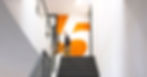 Eine orange Wand mit einer großen weißen Fünf. Im Vordgerund einer lange Treppe mit grauem Teppich. Es ist ein Mann schemenhaft zu erkennen.