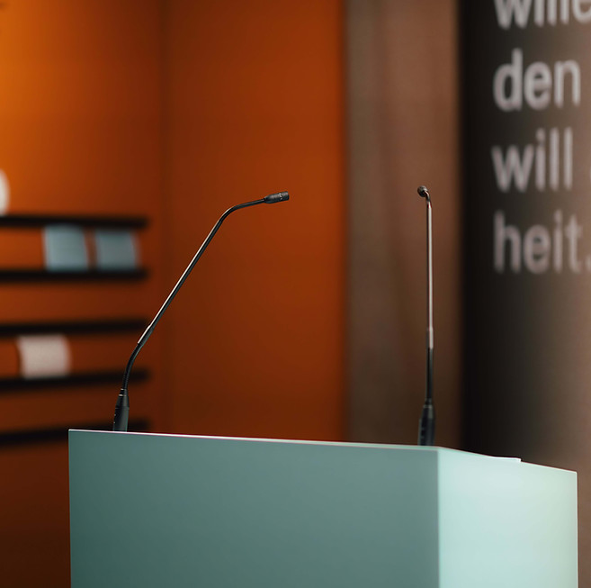 Detailfoto eines hellblauen Rednerpults mit zwei Mikrofonen in einem apricot-farbenen Ausstellungsraum. 