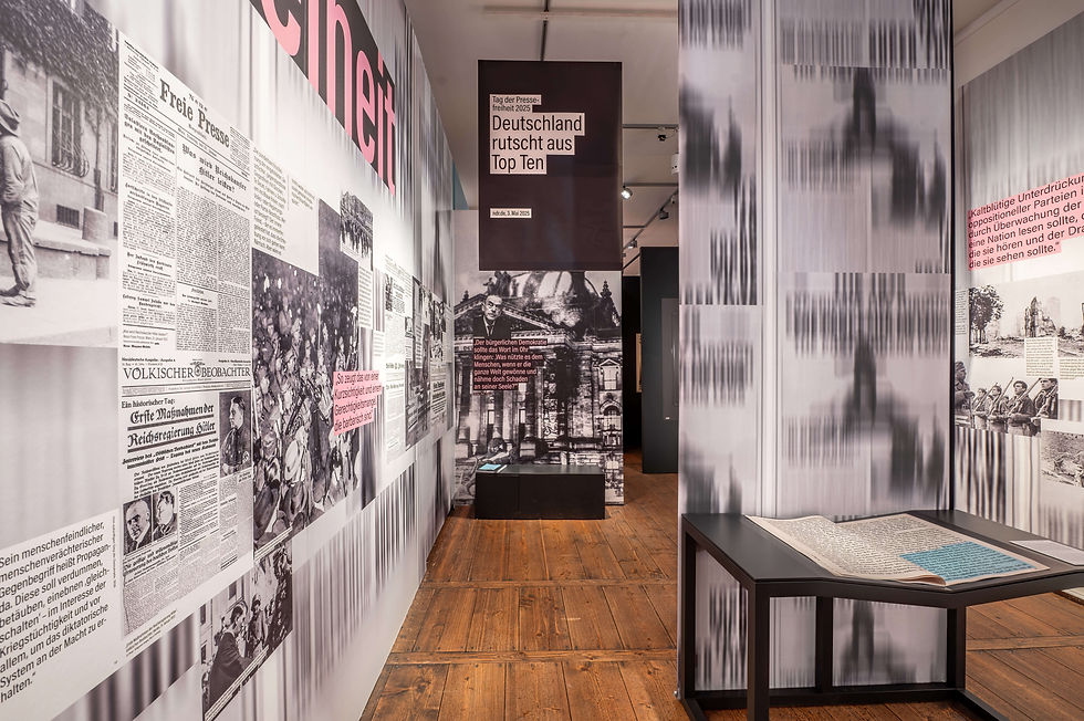 Ein Ausstellungsraum, der kompett mit Zeitungsbahnen, großen Überschriften und Zeitungsartikeln tapeziert ist. Von der Decke hängen einzelne Banner mit Überschriften. In der Mitte ein Sockel mit einer großen Zeitung.