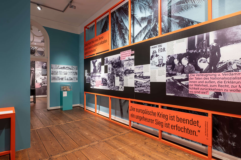 Ein großes Billboard-Gerüst in orange, auf dem zahlreiche Texttafeln und Großabbildungen befestigt sind. Im Hintergrund eine blaue Palmentapete.