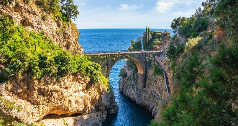 Arched stone bridge over a narrow rocky canal with the blue sea in the background and lush vegetation on the slopes in Furore