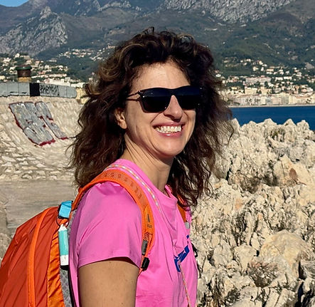 A lady posing for a photo beside some rocky terrain in the bright sunshine with montains in the background