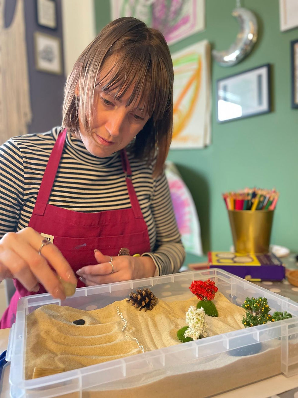 Woman in a red apron arranges a sand tray miniature with tiny trees and a pinecone in a colorful craft room