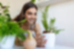 Woman smiling while tending to a potted fern plant indoors, surrounded by fresh green houseplants.
