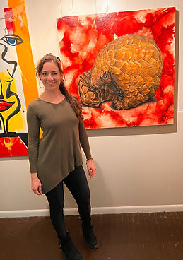 Local artist Katie Pena standing in front of her painting of a pangolin on display in a fine art gallery in Raleigh, NC.