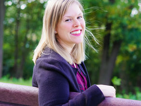Woman smiling, sitting on a bench in a park with green trees in the background. She's wearing a dark jacket, and the mood is cheerful.