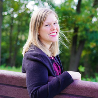 Woman smiling, sitting on a bench in a park with green trees in the background. She's wearing a dark jacket, and the mood is cheerful.