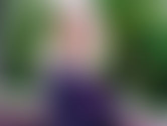 Woman smiling, sitting on a bench in a park with green trees in the background. She's wearing a dark jacket, and the mood is cheerful.