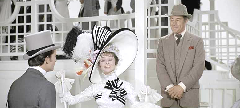 The aristocrat Freddy Eynsford-Hill (Jeremy Brett, left) admires the recently refined Cockney girl Eliza Dolittle (Audrey Hepburn, center), while a proud Professor Higgins (Rex Harrison, right) looks on. Image courtesy of IMDb.com.