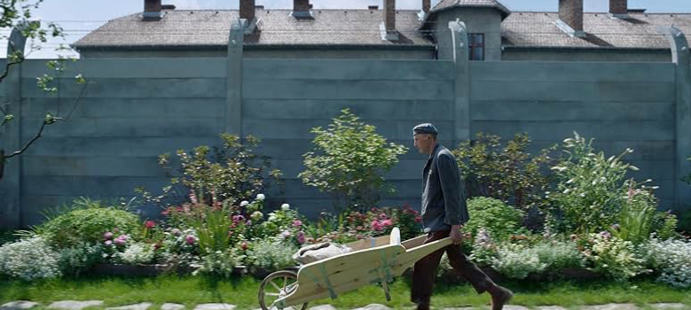 A man pushes a wheelbarrow through a seemingly peaceful and innocent garden, while unseen prisoners are tortured inside the concentration camp just behind the wall. Image courtesy of IMDb.com.