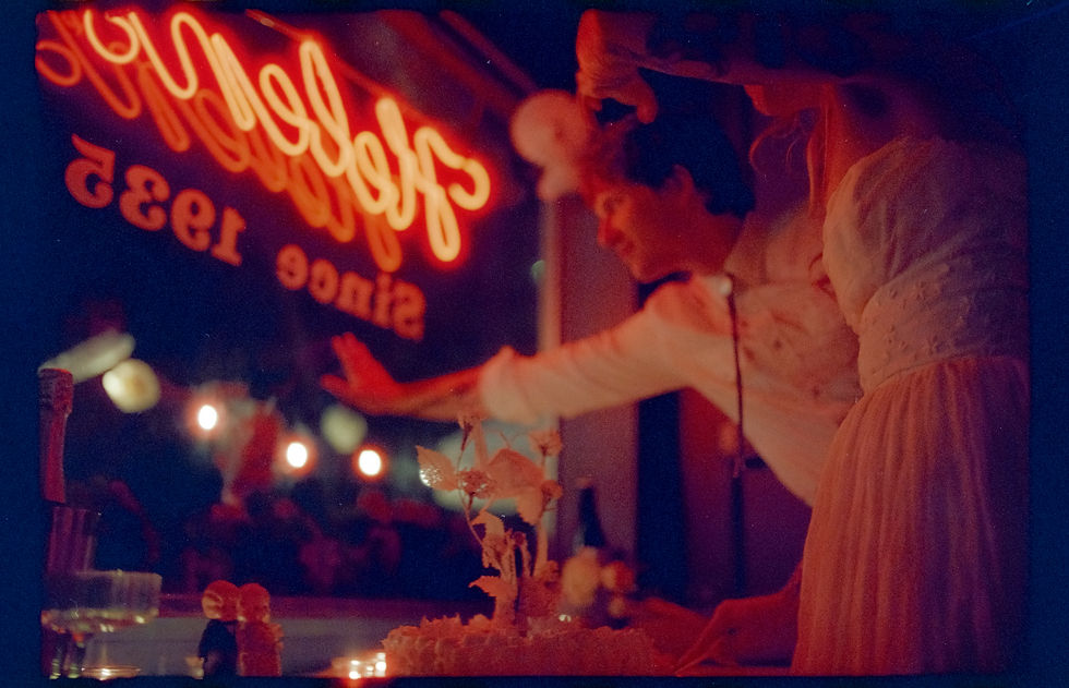 a moody, neon-lit red scene in front of the bar window, a couple waves outside to people sitting on the porch