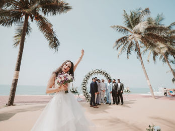 A bright, wide-angle beach wedding photograph taken in Udupi captures a jubilant bride in the foreground, wearing a layered white tulle gown and holding a purple and white bouquet. She celebrates with one arm raised against a clear blue sky. In the middle ground, the groom and five groomsmen in various suits stand before a circular floral arch adorned with white roses. The scene is framed by tall, leaning coconut palms, with the serene Arabian Sea visible in the distance. The warm, sandy tones of the coastal venue and the high-angle perspective emphasize the joyful, expansive atmosphere of the celebration. The lighting is bright and natural, highlighting the vibrant coastal setting and the energetic emotion of the wedding day.