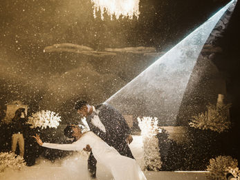 A dramatic, vertical photograph captures a cinematic first dance during a Christian wedding in Kochi, Kerala. The groom, in a dark navy tuxedo, dips the bride, who wears a sophisticated long-sleeved white gown. They are center stage, enveloped in a thick layer of low-lying fog and shimmering confetti that resembles falling snow. A powerful white spotlight cuts through the dark venue, illuminating the couple and the swirling particles. Above them, a large, ornate chandelier of white flowers hangs from the ceiling. The warm, golden-toned lighting and the ethereal atmosphere create a magical, high-energy moment of celebration. White floral arrangements and a photographer in the background frame the stage, emphasizing the grandeur of the event.