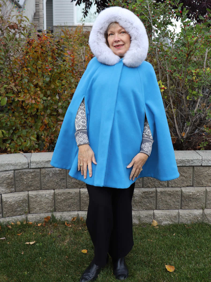 A woman wearing an elegant, cerulean blue cape with a blue fox fur trimmed hood. The hood is up.