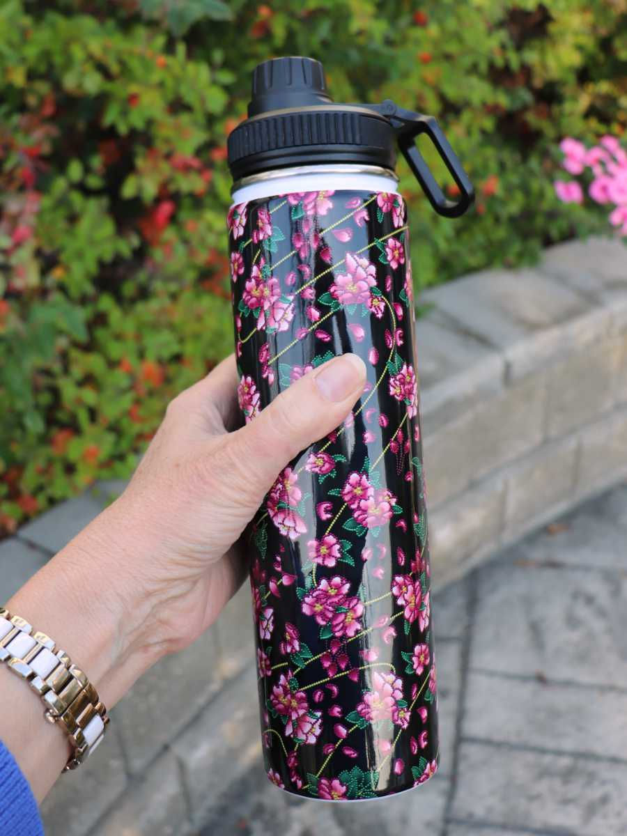Thumbnail: A hand holding a black and pink, stainless steel, screw top water bottle patterned with pink, wild roses with beaded edges