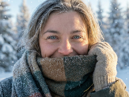 Primer plano de una mujer sonriente protegiéndose del frío invernal con una bufanda gruesa de cuadros y guantes de lana, con escarcha visible en su cabello y pestañas en un paisaje nevado.