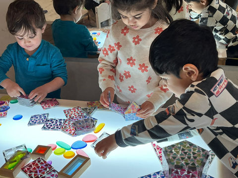 Three children working at a light table with colorful translucent tiles and shapes