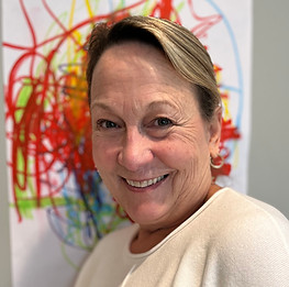 Smiling woman with short light brown hair wearing a cream-colored top, standing in front of a colorful abstract drawing.