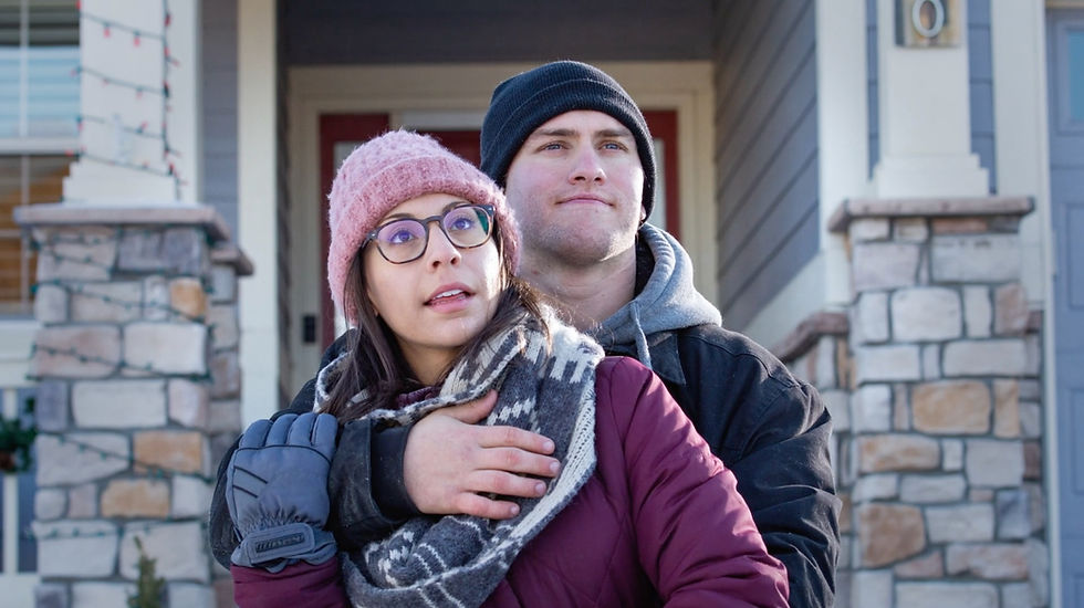 Couple in winter attire stand on porch; woman in pink hat, man in black beanie. Cozy mood, stone columns in the background.