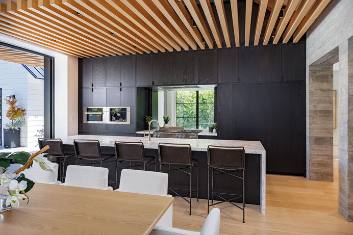 Modern kitchen with a large island, white cabinets, stainless steel appliances, and granite countertops opens to a dining room with a table, chairs, and a light fixture.  Real estate photography by Adam Dubich Photography, Orange County, California.