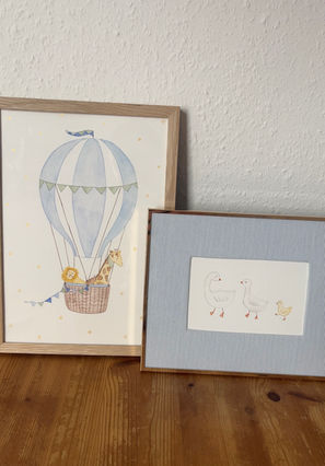 Das Bild zeigt zwei gerahmte Aquarellbilder. Auf dem einen ist ein Heißluftballon zu sehen. Im Korb sitzen eine Giraffe, ein Löwe und ein Hase. Das zweite Bild zeigt zwei erwachsene Gänse und eine Baby Gans.