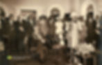 Sir Alexander Bustamante sits front and center in a suit and sash, while Gladys Maud Bustamante speaks at a podium with microphones. They are surrounded by formally dressed people in a room with arched windows and floral decorations.