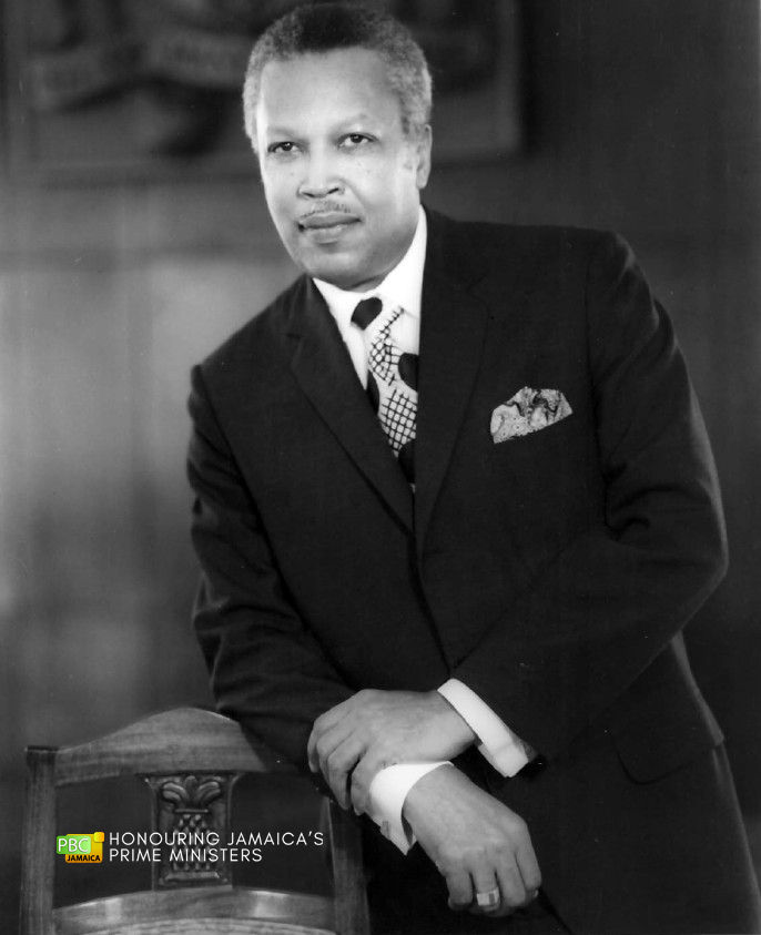 Monochrome image of Hugh Shearer in a dark suit and patterned tie with large polka dots and cross-hatching, leaning on a chair with a confident expression.