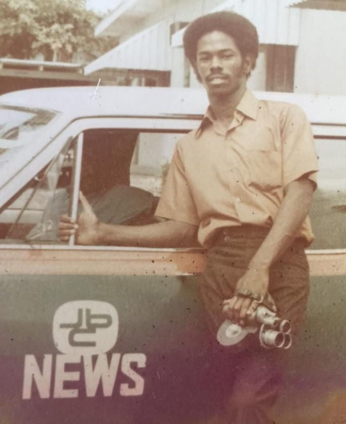 Errol Russell, dressed in a short-sleeved shirt and dark trousers, stands beside a JBC News vehicle holding a vintage film camera, with one hand resting on the car door.