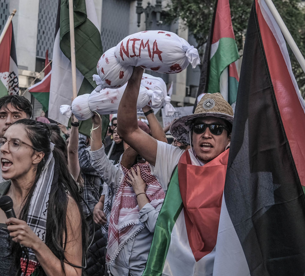Woman in a keffiyeh crying out while carrying a white wrapped bundle representing children lost in Gaza