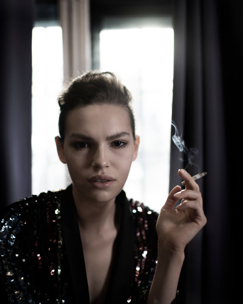 Female model in black sequin blazer holding cigarette in dark dramatic editorial portrait, I.M.G Modeling by The Content Campus