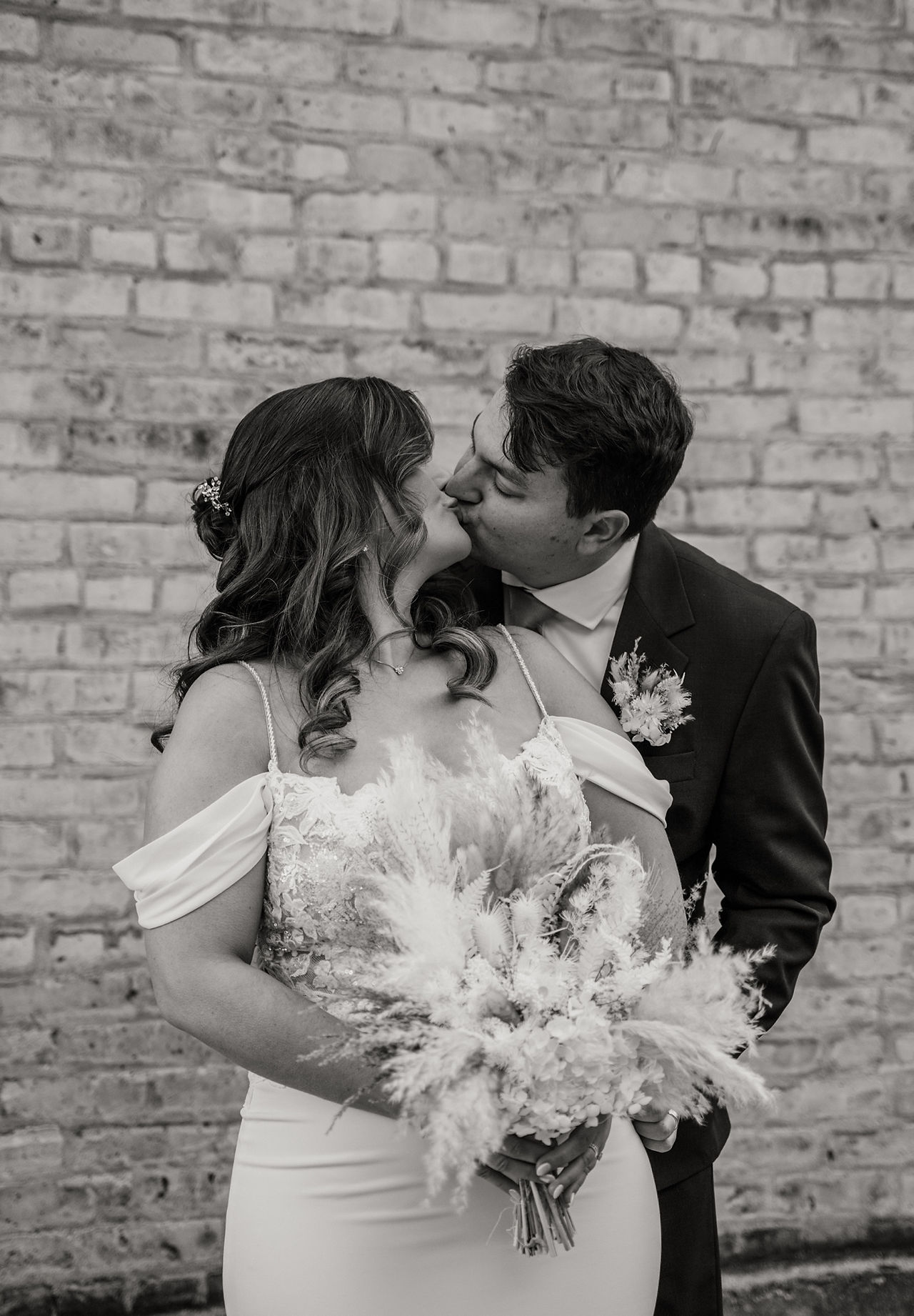 Black and white photo of a bride and groom kissing while standing in front of a brick wall