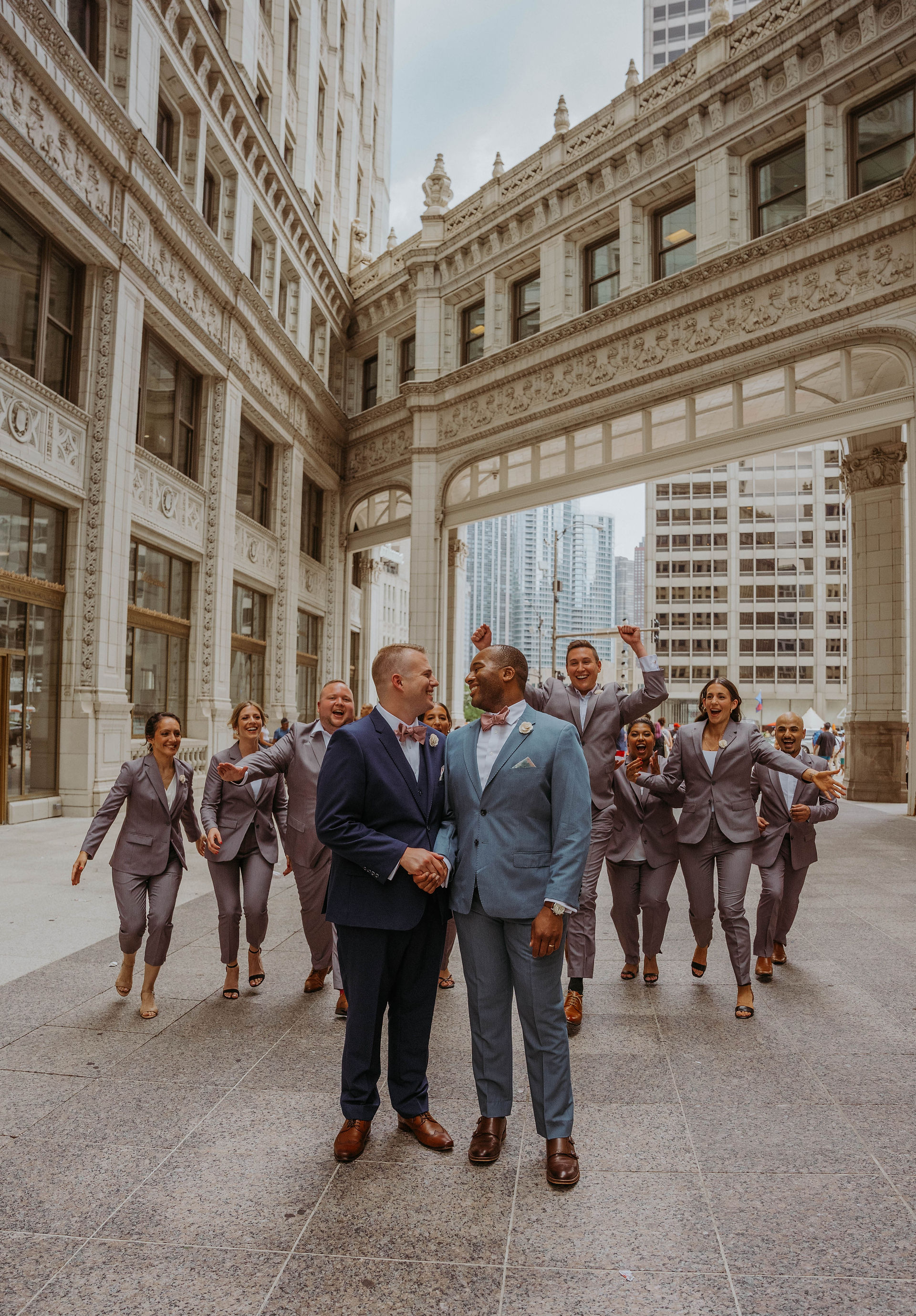 Grooms look at each other as their bridesmaids and groomsmen in grey suits run up behind them. They are in the large outdoor corridor of the Wrigley Building in downtown Chicago. 