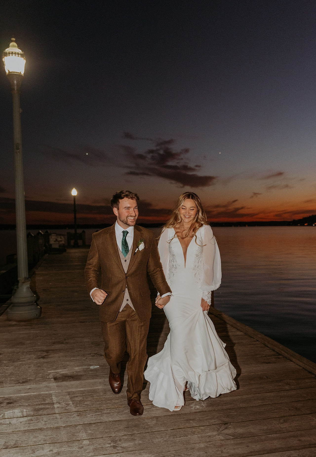 A bride and groom hold hands as they walk along a pier in Lake Geneva while the sun sets behind them over the water