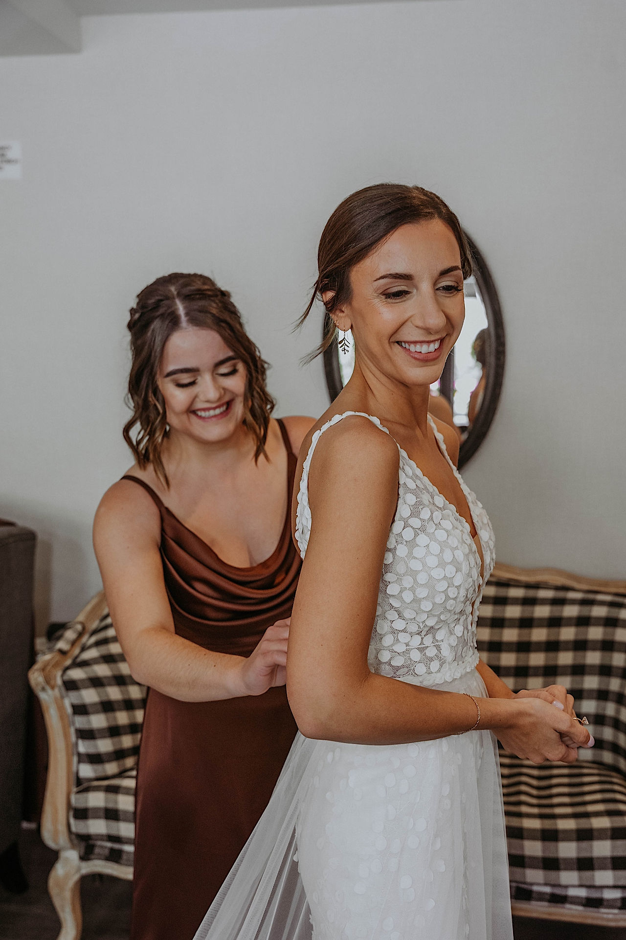 Bride smiles as bridesmaid helps zip up her lace wedding dress while getting ready for wedding in a farm style living room. 