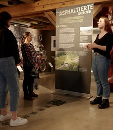 Junge Frau erklärt Besucherinnen eine Ausstellungstafel mit dem Titel „Asphaltierte Grenzen“ im PS.SPEICHER.