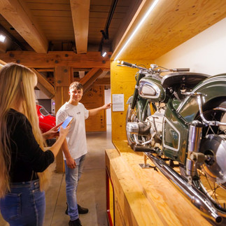 Ein junger Mann zeigt auf die Beschreibung eines Oldtimer-Motorrads in einem Museum, während eine junge Frau ein Foto davon macht während der Erlebnistour durch den PS.SPEICHER