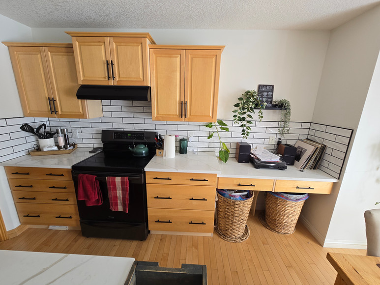 BEFORE photo showing the cooking and workspace area of the kitchen, with light cabinets, original hardware, and black appliances. White countertops and subway tile backsplash with dark grout are already in place.