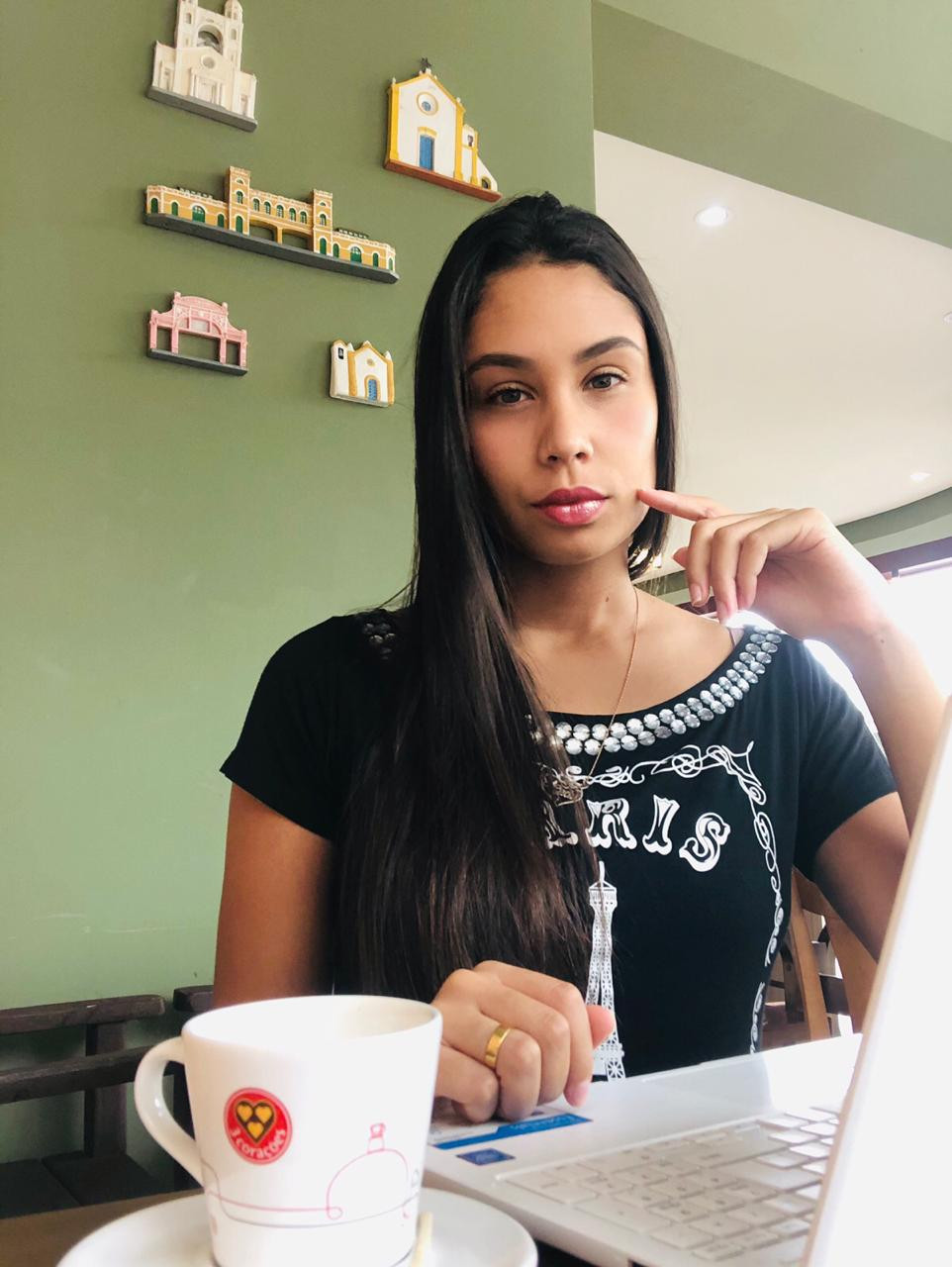 Na foto está a Karine, uma moça jovem sentada em uma cafeteria com o seu notebook e uma xícara de café. Ela trabalha como Redatora Freelancer.