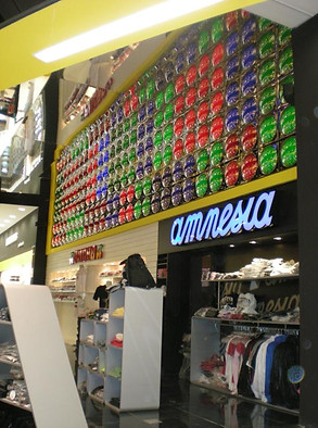 A shop of t-shirts and caps in the mall, on one wall there is an impressive display made up by colourfull glass balls with different colours inside. The store has white walls and yellow trim. There's some soccer shirts displayed at ground level. It feels modern but casual.