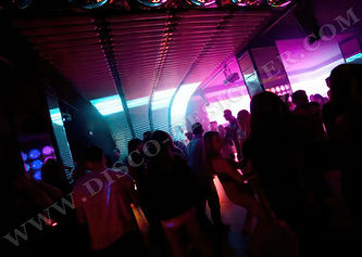 photo of a party in a night club, people dancing and having fun, green lights, neon lights on the ceiling, modern architecture, large windows with a city view, white walls, black leather sofa, a girl in a red dress standing near a table, a big LED screen showing music from the DJ stage, green lights on the floor