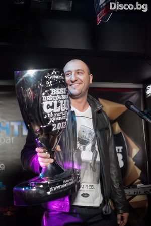Photo of a smiling man in a leather jacket holding a large trophy cup with text "best club in Bulgaria" on it, photorealistic, on stage near the microphone under studio lighting.
